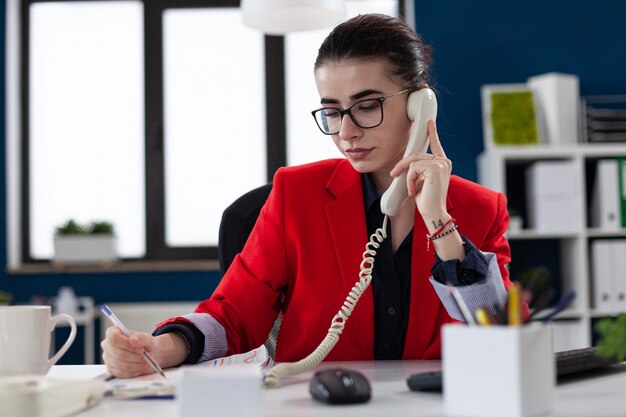 businesswoman taking notes on clipboard sitting at desk in corporate office while taking