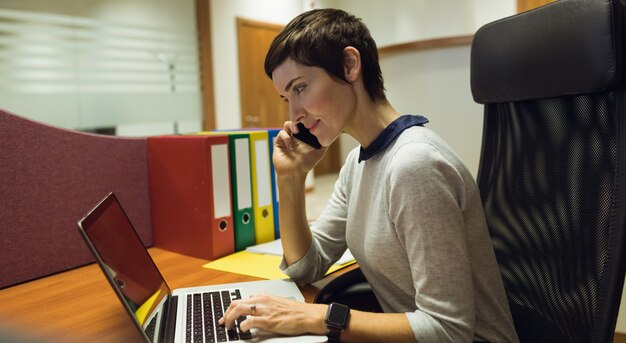 Деловая женщина разговаривает по мобильному телефону, используя ноутбук в офисе