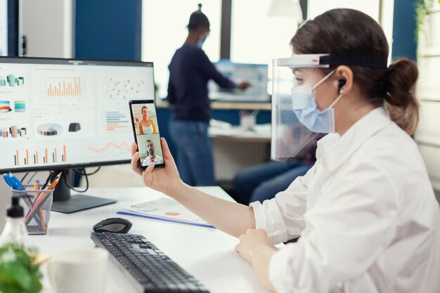 Деловая женщина с помощью беспроводных наушников во время онлайн-конференции на смартфоне в маске на рабочем месте. Многонациональные коллеги, соблюдающие социальную дистанцию в бизнесе во время глобальной пандемии