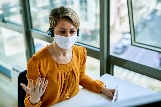 Donna d'affari con auricolare che comunica mentre lavora in ufficio e indossa una maschera facciale