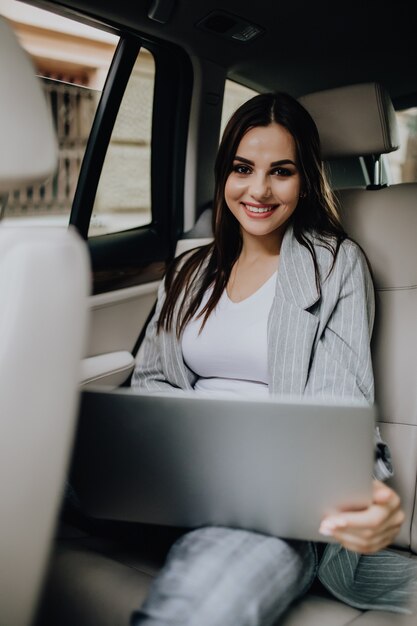 Businesswoman with laptop receiving a phone call on the backseat of a car. Female entrepreneur working during travelling to office in a car.