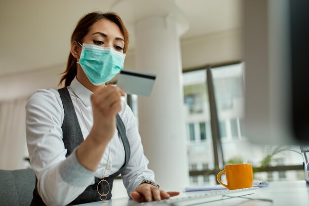 Businesswoman with protective face mask using computer and credit card while ebanking in the office