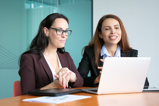 Businesswomen having video talk to partners, sitting at open laptop, looking at display and smiling