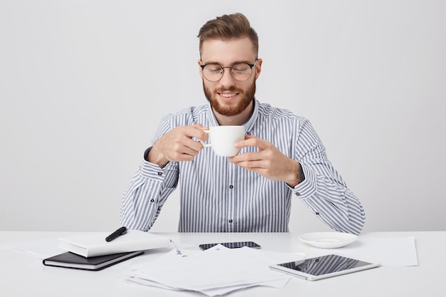 厚いひげを持つ忙しい魅力的な男はコーヒーブレークを持っている、職場に座っている、ドキュメントを扱う