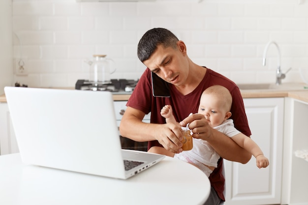 Busy brunette male wearing maroon casual style t shirt sitting at table in kitchen feeding his infant daughter with fruit puree, talking via cell phone with business partner.