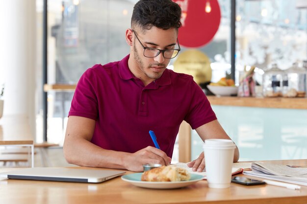 Busy businessman wears glasses and t shirt, writes down information in notepad, prepares ideas for startup project, drinks coffee and eats croissant, poses in bistro against blurred wall.