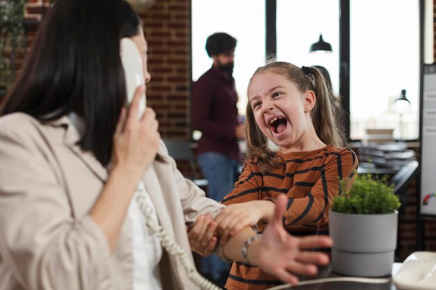 Busy businesswoman trying to talk on telephone while screaming and annoying daughter disturbs her in company office. Young mother working and sitting at desk while little girl interrupts her.