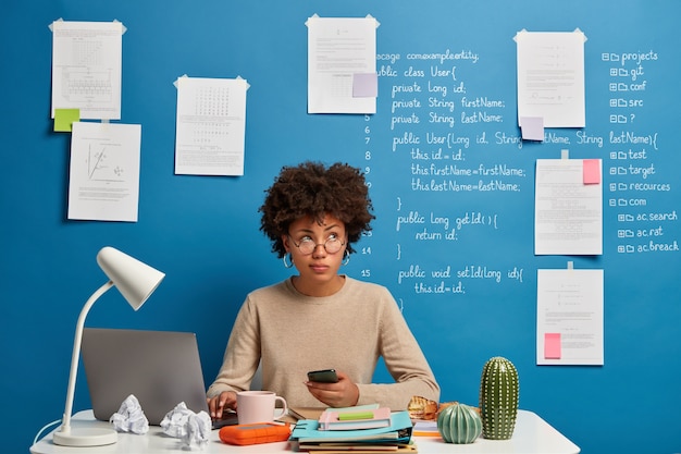 Busy curly Afro woman works from home, uses laptop and smartphone at workplace, checks newsfeed, poses at white desk with folders and notepads.