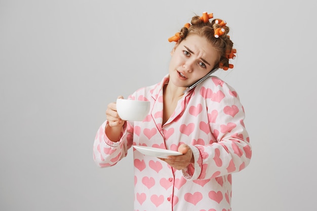 Free photo busy cute girl in hair curlers and pajamas, drinking coffee and talking on phone