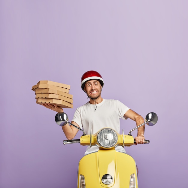 Busy deliveryman driving yellow scooter while holding pizza boxes