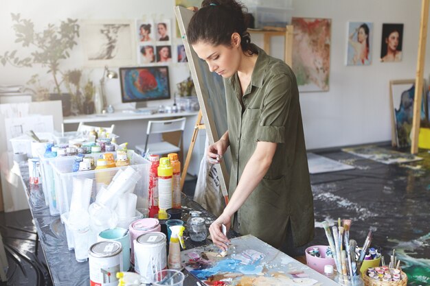 Busy female painter taking oil colors while standing near table with oils, working in art studio, going to draw sea landscape or portrait. Attractive young woman working on canvas at workshop