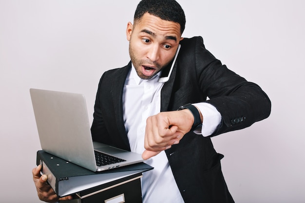 Busy handsome guy in white shirt and black jacket talking on phone, holding folders, laptop, looks astonished at watch. Being late, meeting, office worker, career.