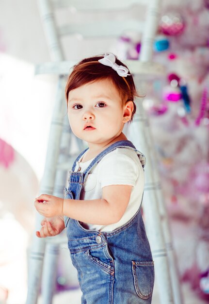 Busy little girl in jeans jumpers stands before tall white chair 