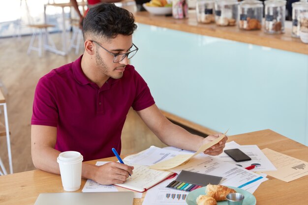 Busy male expert studies marketing issues, surronded with documents, learns graphhics and diagrams, uses stickes and notepad for noting information, spends lunch time in cafeteria or coffee shop