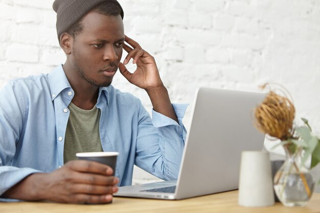 Busy male with dark skin looking seriously in laptop computer while reading news online, keeping paper cup with coffee, resting in cafeteria. Handsome male reading electronic book on computer