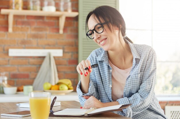 実業家の忙しい朝。うれしそうな笑顔の女性は、彼女の作業スケジュールをらせん状の日記に書いて、肯定的な感情を表現します、