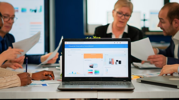 Busy multicultural diverse employees analysing annual financial statistics sitting at conference desk in back of laptop holding documents searching business solutions. Business team working in company