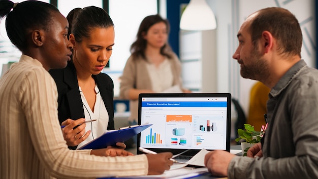 Busy multicultural, diverse employees analysing annual financial statistics sitting at desk in front of laptop holding documents searching business solutions. Team of businesspeople working in company