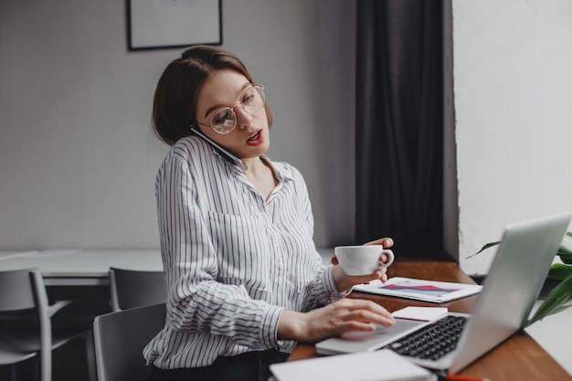 Busy office worker talking on phone and working in laptop, holding cup of tea.