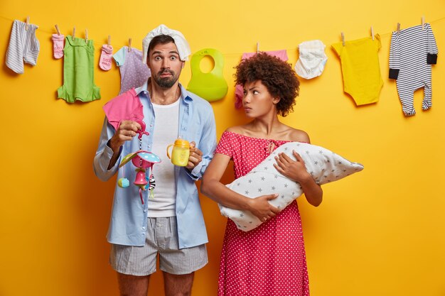 Busy parents nursing baby. Young couple experience parenthood. Newborn infant in hands of caring mother. Shocked father holds feeding bottle and mobile, diaper on head. Parenting, family concept