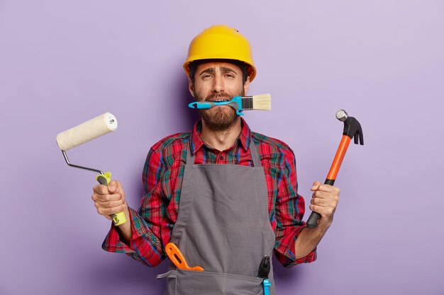 Busy repairman holds building tools, does repairing at home, wears yellow hardhat, apron, stands indoor.