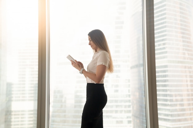 Free photo busy serious businesswoman using digital tablet, standing near big window