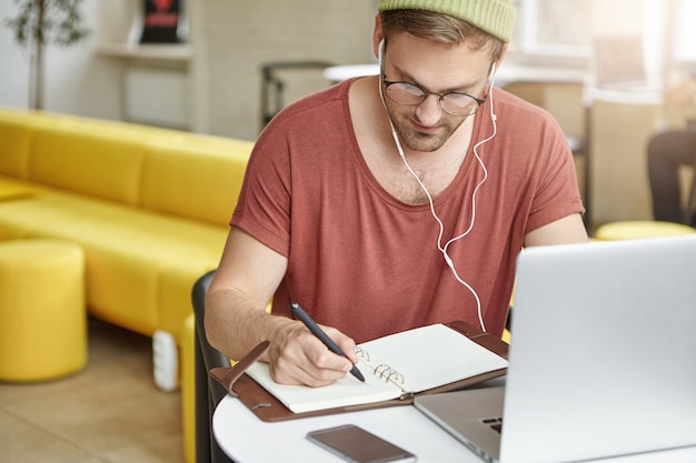 Free photo busy student wears round glasses and hat, write notes in notebook,