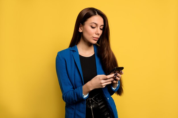 Busy stylish business woman with long dark hair using smartphone over isolated wall. European woman scrolling phone over yellow wall