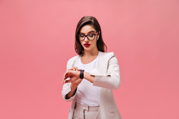Busy woman in beige jacket looks at her hand watch. Brunette with red bright lips in glasses and white headphones posing on isolated background.