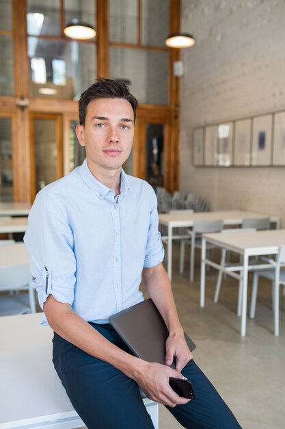 Busy young attractive smiling man sitting in co-working open office, holding laptop