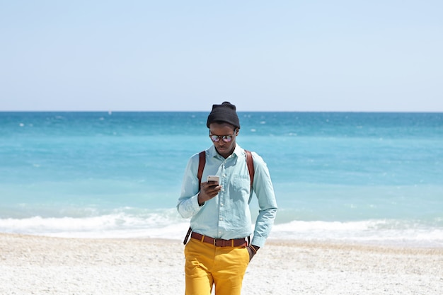 Busy young dark-skinned European man wearing fashionable trendy clothing and backpack staying online even during vacations, using mobile phone on beach, ignoring all beauties that surround him