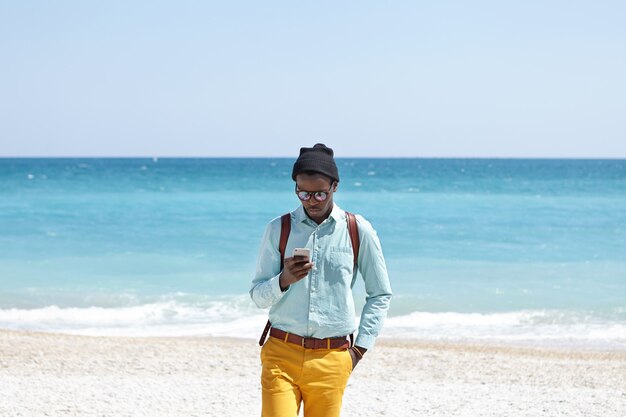 Busy young dark-skinned European man wearing fashionable trendy clothing and backpack staying online even during vacations, using mobile phone on beach, ignoring all beauties that surround him