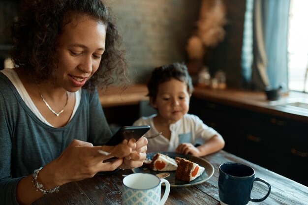 Busy young single mother using cell phone for distant work, writing posts on social network accounts