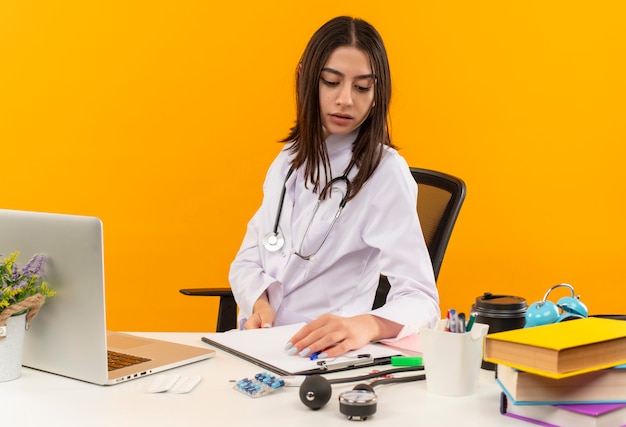 Foto gratuita occupato giovane donna medico in camice bianco con stetoscopio seduto al tavolo con laptop e documenti sulla parete arancione