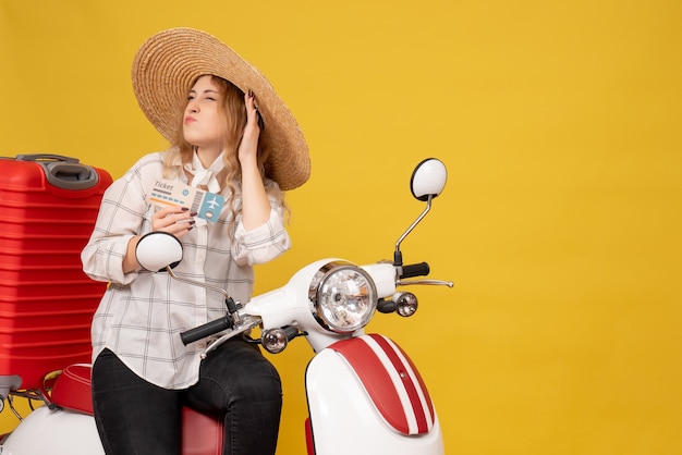 Busy young woman wearing hat and sitting on motorcycle and holding ticket listening the last gossip on yellow 