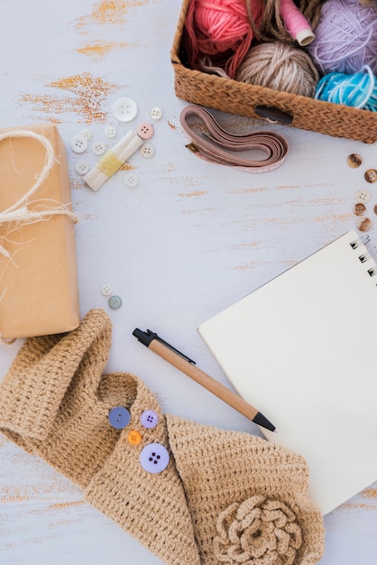 Buttons; wrapped gift box; measuring tape; pen; knitted scarf and spiral notepad on white wooden table