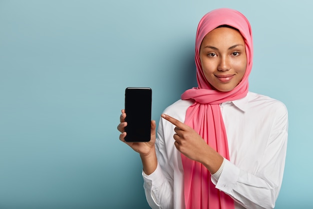 Free photo buy this device! pleased dark skinned woman in pink veil, points at smart phone