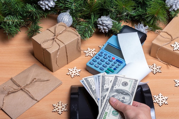 Покупка подарков на рождество и новый год - рука кассира, держащая наличные доллары над кассовым аппаратом, для оплаты рождественских подарков на прилавке с украшенными еловыми ветками. рождественская распродажа