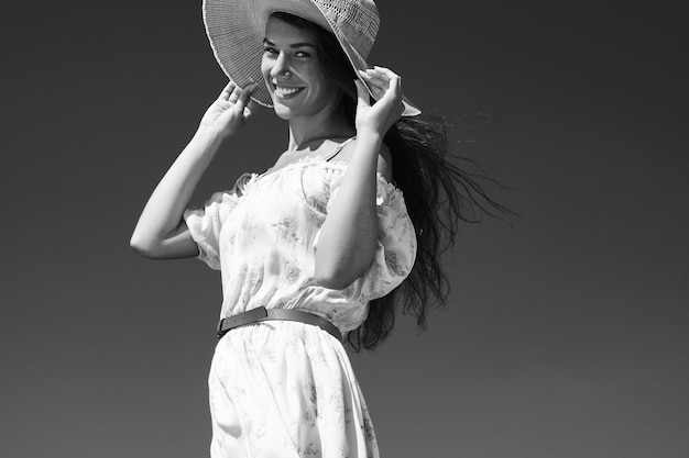 바람에 날리는 여름 모자를 잡은 여자의 Bw 초상화는 꽃무늬 드레스를 입는다