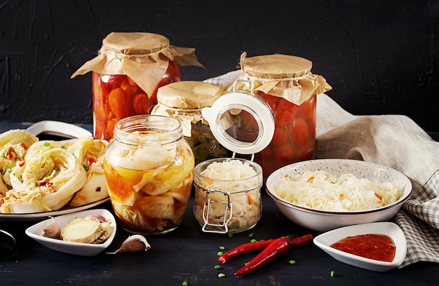 Cabbage kimchi, tomatoes marinated, sauerkraut sour glass jars over rustic kitchen table.