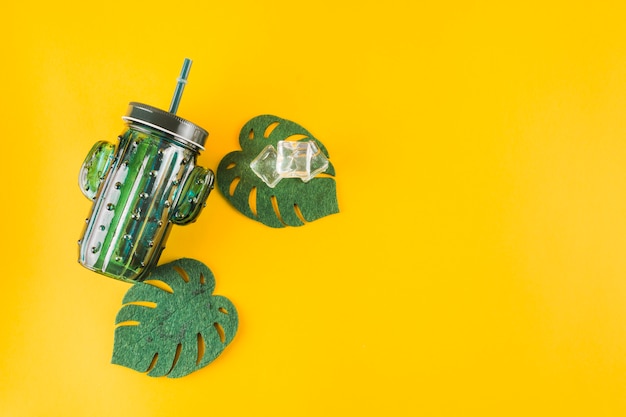 Cactus shape jar with ice cubes and artificial monstera leaves on yellow background