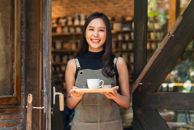 Cafe female owner smiling with positive happiness ready to open for customerasian female woman with apron hand present tray and coffee cup warm welcome condident standing at entrance of coffeeshop