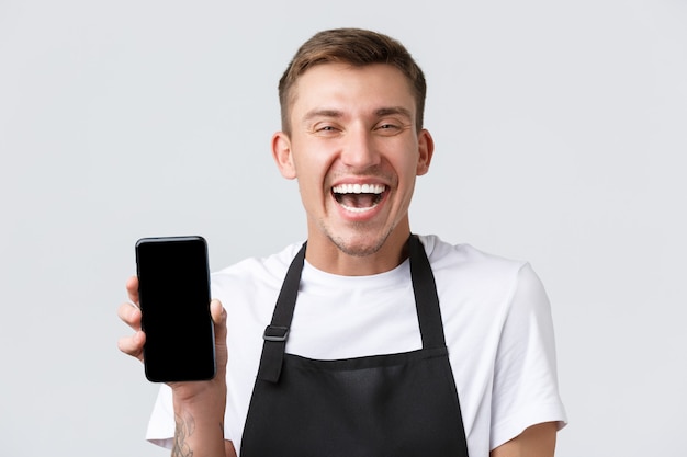 Cafe and restaurants, coffee shop owners and retail concept. Cheerful salesman in black apron showing smartphone app and laughing happy, inform clients new application, delivery option