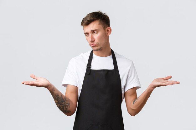 Cafe and restaurants, coffee shop owners and retail concept. Disappointed gloomy waiter, salesman in black apron sighing upset, showing two variants on palms and looking sad at left hand