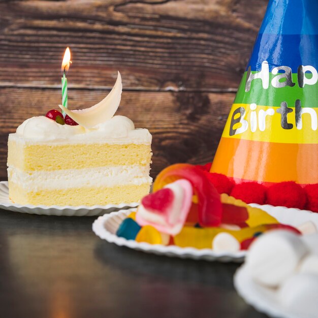 Cake slice; candies; marshmallow and birthday hat on table