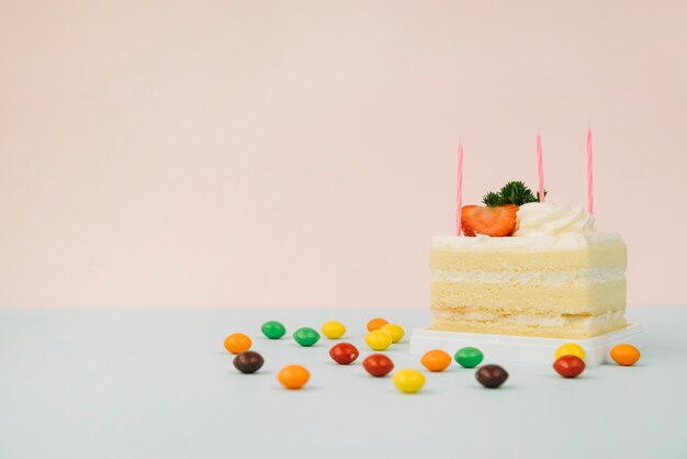 Cake slice with candles and candies on table against pink background