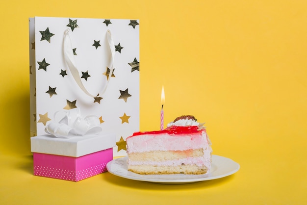 Cake slice with lighted candle; star shape shopping bag; and gift box on yellow background