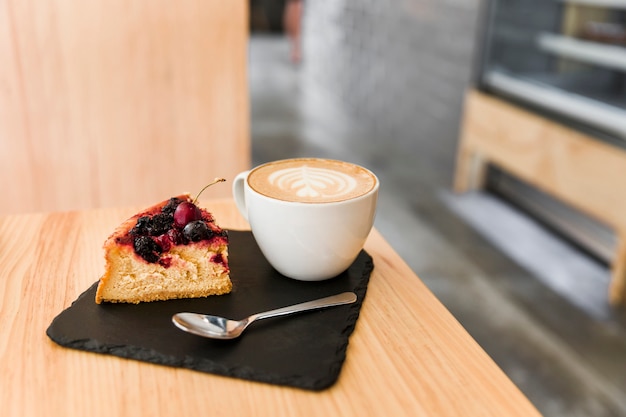 木製テーブル上のシェールボード上にスプーンとカプチーノのコーヒーを入れたケーキスライス