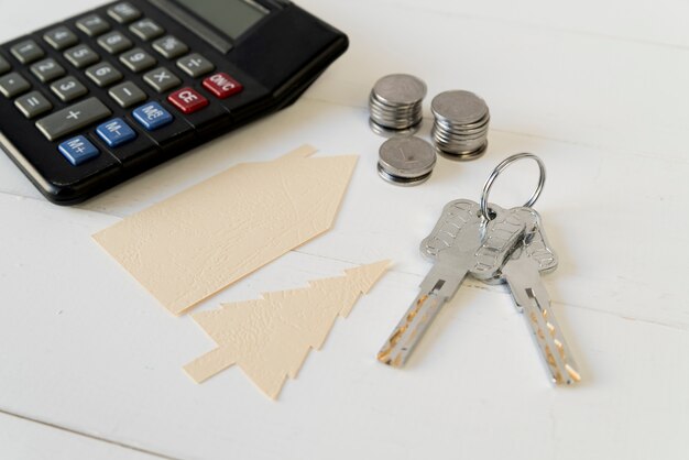 Calculator; coins stack; keys with house and tree paper cutout on white wooden table