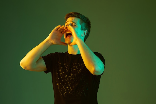 Calling somebody, screaming. Caucasian man's portrait isolated on green studio background in neon light. Beautiful male model in black shirt. Concept of human emotions, facial expression, sales, ad.
